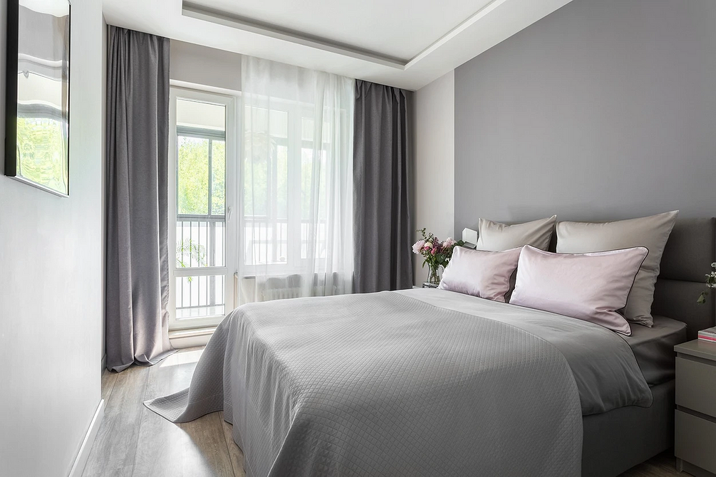 Затишна спальня з сірим ліжком, килимом та декоративними подушками в нейтральних тонах.